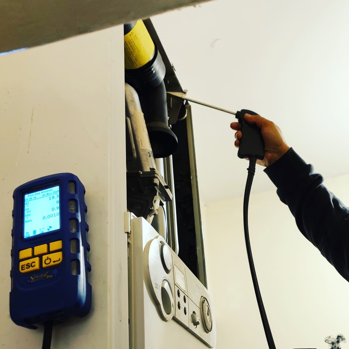 A hand holding a flue gas analysis probe inside a boiler flue, with a blue Sprint Pro analyser reading oxygen and carbon monoxide levels on the wall beside it.